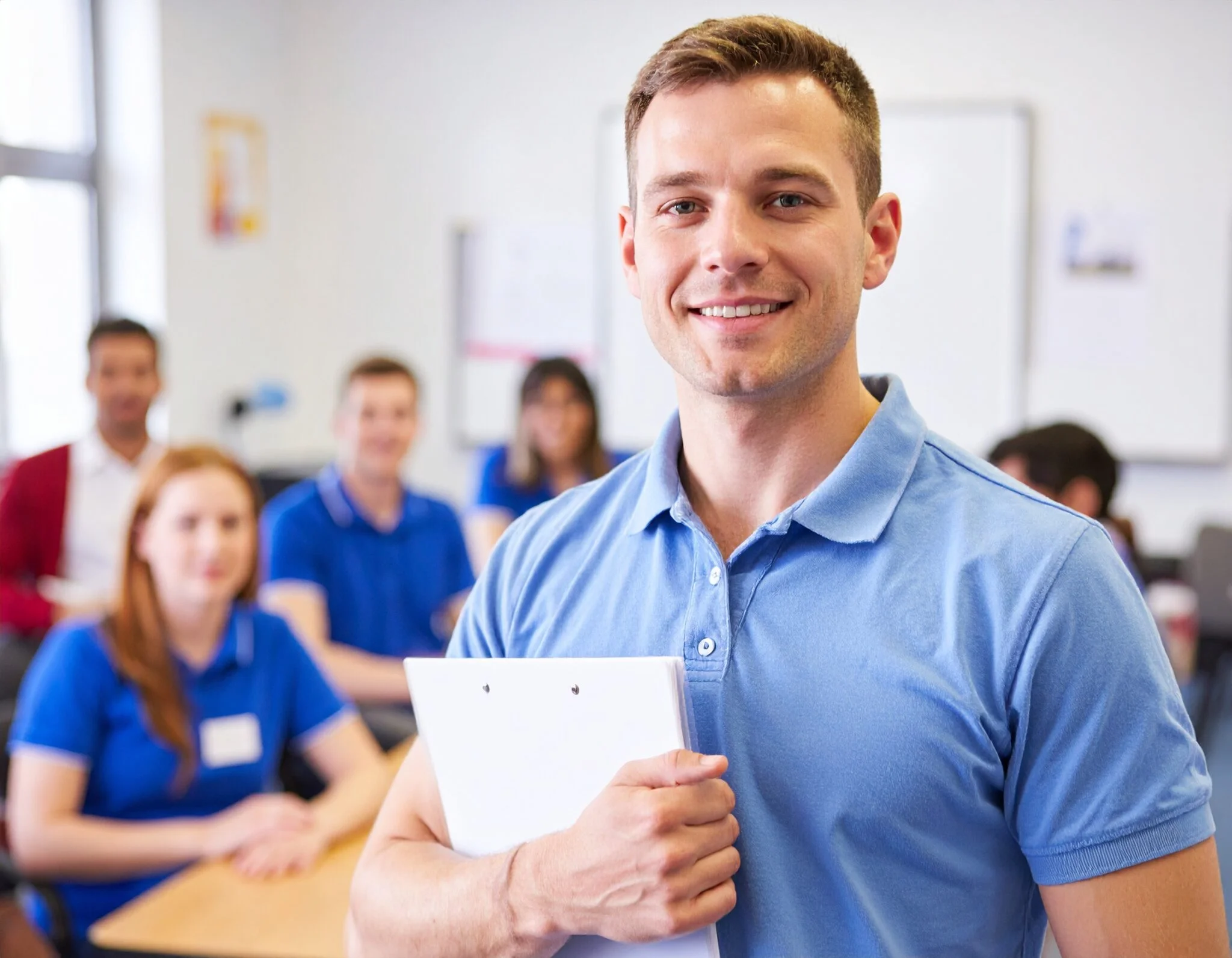 Confident male first aid instructor with clipboard in training classroom environment.
