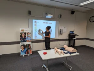 Classroom first aid training session teaching DRSABCD protocol with CPR mannequins and projection screen display.