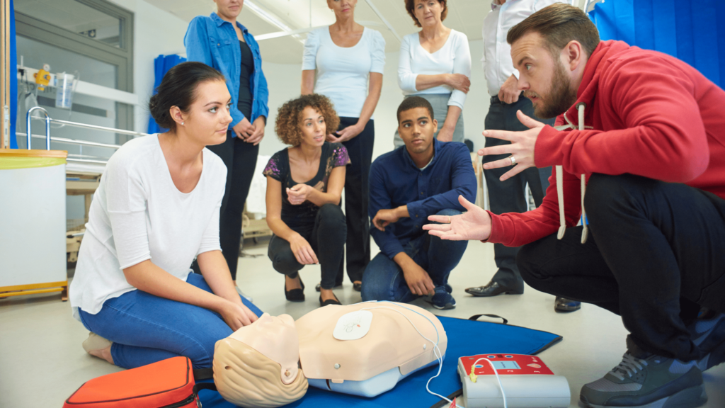 First aid instructor teaching a student how to use a defibrillator on a CPR manikin during a group training session
